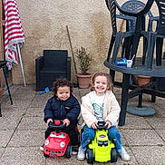 Kayden a rejoint le concours — aidez-le/la à gagner de superbes lots ! child, toy_car, outdoor, grass, pavement, plastic_chair, umbrella, plant_pot, smiling, jacket, happy, play, fun, backyard, green, red, person, casual, chair_stack, toy