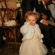 Eloïse participe au concours pour gagner de l'argent avec cette photo : toddler, child, curly_hair, white_jacket, cream_dress, smile, indoor, formal_clothing, seated_adults, wooden_chairs, event, flower_decoration, happy, portrait, floor, people, party, cute, dress, celebration