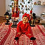 baby, red_clothing, headband, smiling, christmas_tree, nutcracker, ornaments, blanket, holiday, indoor, decorations, floor, lamp, wooden_floor, festive, toy, face, person, child, happy