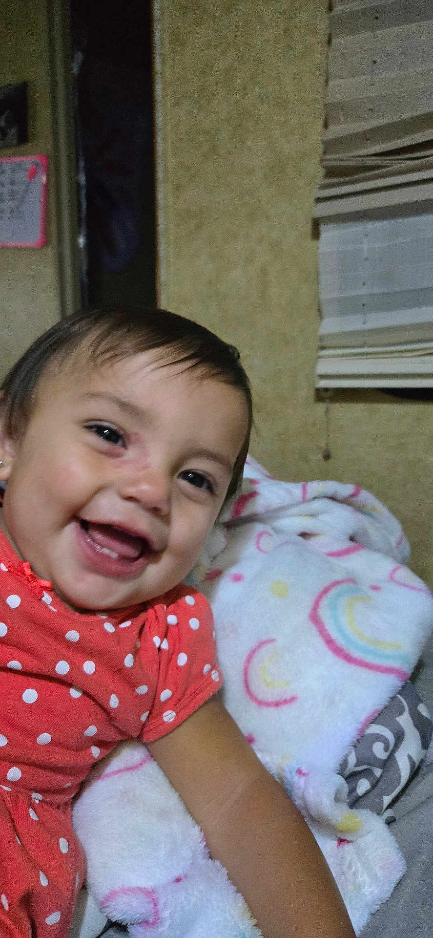 Kapri is registered to the contest to win money with this photo: toddler, child, smiling, face, blanket, polka_dot, red_clothing, indoor, window_blind, happy, cute, baby, portrait, head, skin, person, sitting, fabric, cozy, colorful