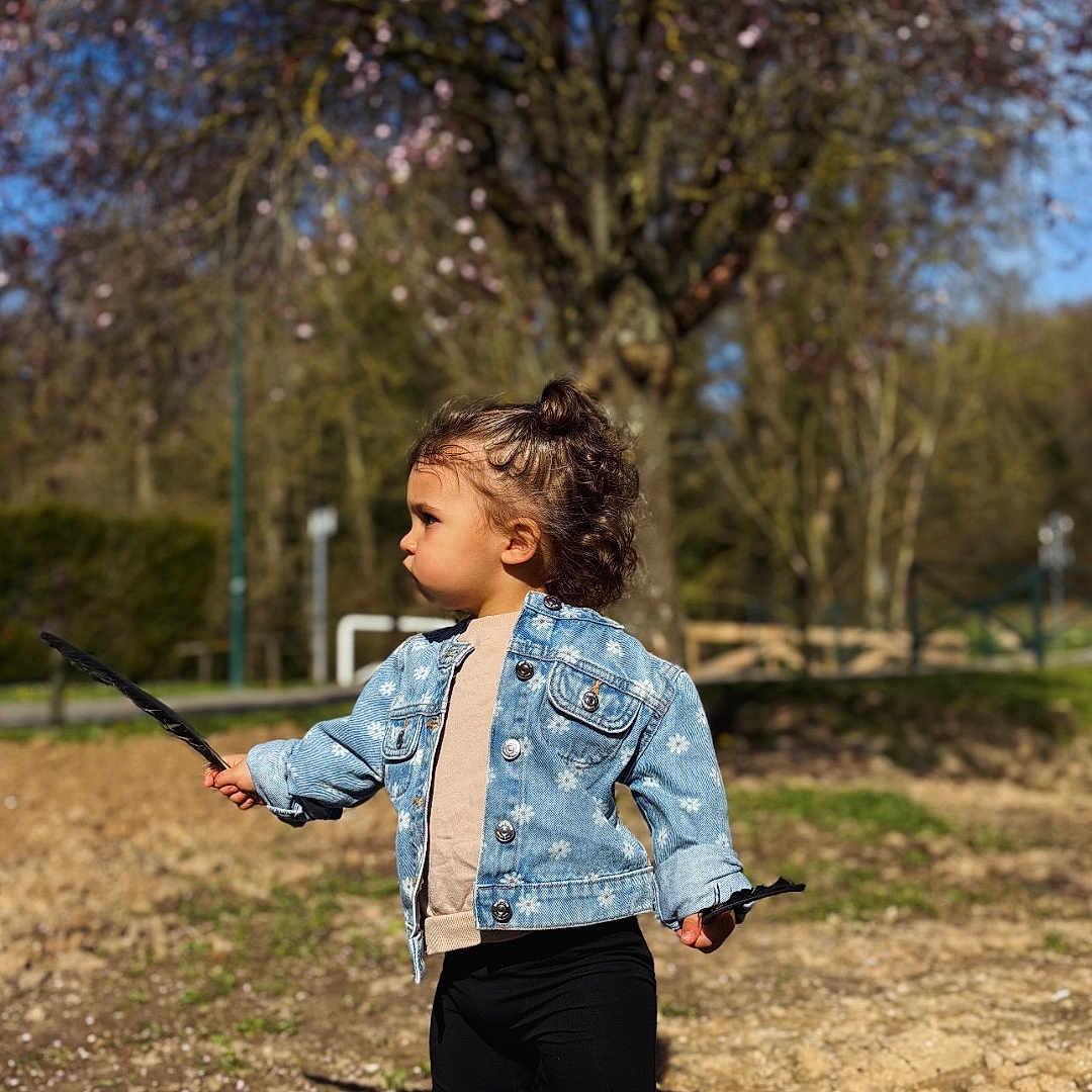 Leynah participe au concours pour gagner de l'argent avec cette photo : toddler, child, denim_jacket, sneakers, sticks, outdoor, spring, blossoming_tree, grass, dirt, sunny, casual_clothing, young_child, nature, park, daytime, curly_hair, standing, playful, determined