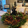 cat, tuxedo_cat, wreath, pine_cones, red_berries, table, dining_room, chair, christmas_tree, holiday_decor, centerpiece, evergreen, indoor, curious, whiskers, black_cat, collar, festive, pine_needles, home_decor
