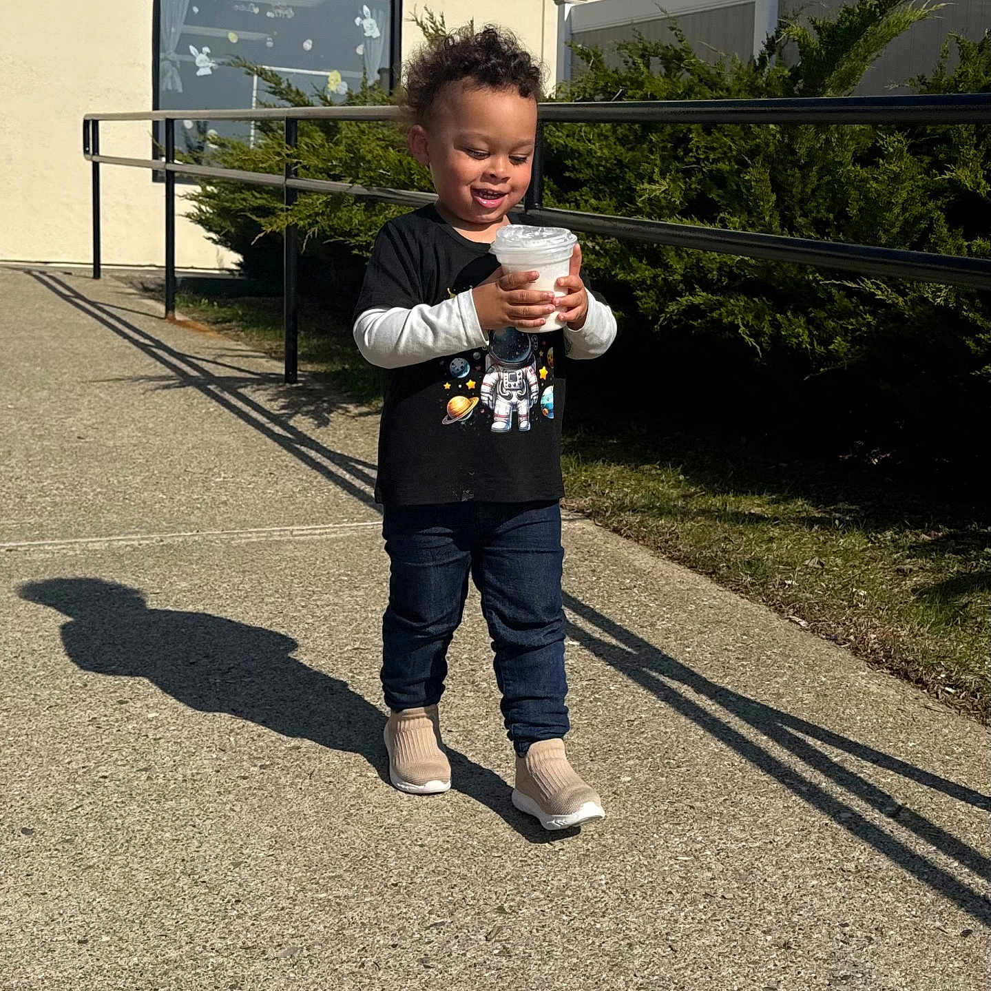 Zidane joined the competition — help win amazing prizes! child, toddler, smiling, outdoor, sidewalk, sunny, holding, cup, black_tshirt, space_theme, jeans, sneakers, shadow, greenery, railing, window, building, curtains, happy, casual_clothing