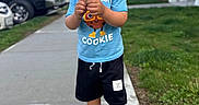 Zidane is registered to the contest to win money with this photo: black_shorts, blue_tshirt, casual_clothing, child, curly_hair, cute, daytime, grass, holding, houses, outdoor, parked_cars, person, portrait, sidewalk, sneakers, suburban, toddler, walking, young_child
