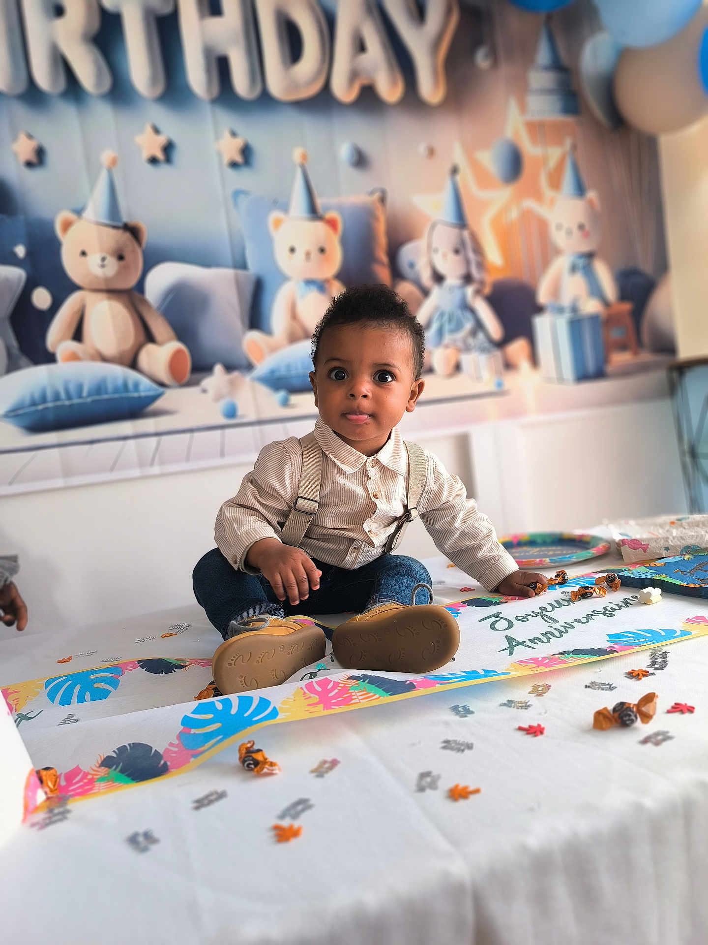 Maëlio participe au concours pour gagner de l'argent avec cette photo : toddler, birthday_party, table, confetti, candy, decorations, plush_toys, teddy_bears, party_hats, child, curious, sitting, clothing, suspenders, jeans, indoor, celebration, smile, face, cute
