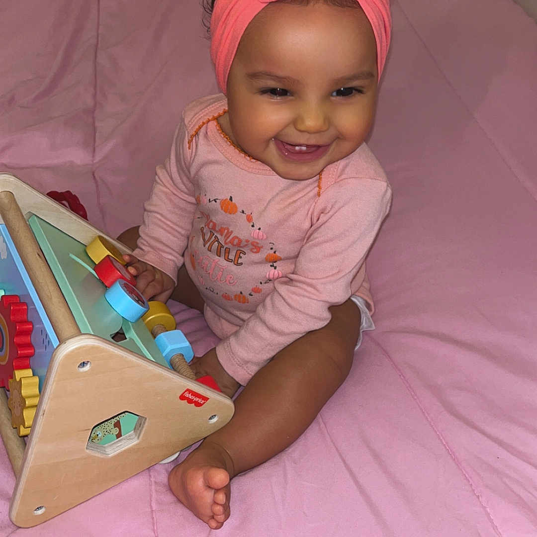 Xymariana is registered to the contest to win money with this photo: baby, bed, blanket, child, clothing, cute, face, foot, girl, happy, headband, indoors, infant, pink, play, room, sitting, smiling, toy, wooden_toy