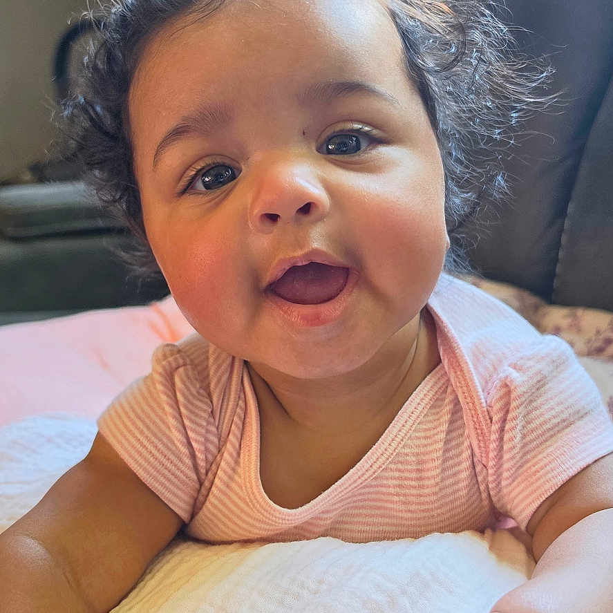 Quinn joined the competition — help win amazing prizes! baby, child, curly_hair, smiling, cushion, pink_clothing, indoors, natural_light, window, happy, face, skin, person, infant, toddler, cute, portrait, soft_texture, home, cozy