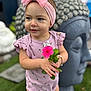 toddler, child, flower, pink_bow, pink_outfit, bracelet, buddha_statue, garden, greenery, grass, cute, portrait, smiling, headband, outdoor, person, nature, statue, happy, baby