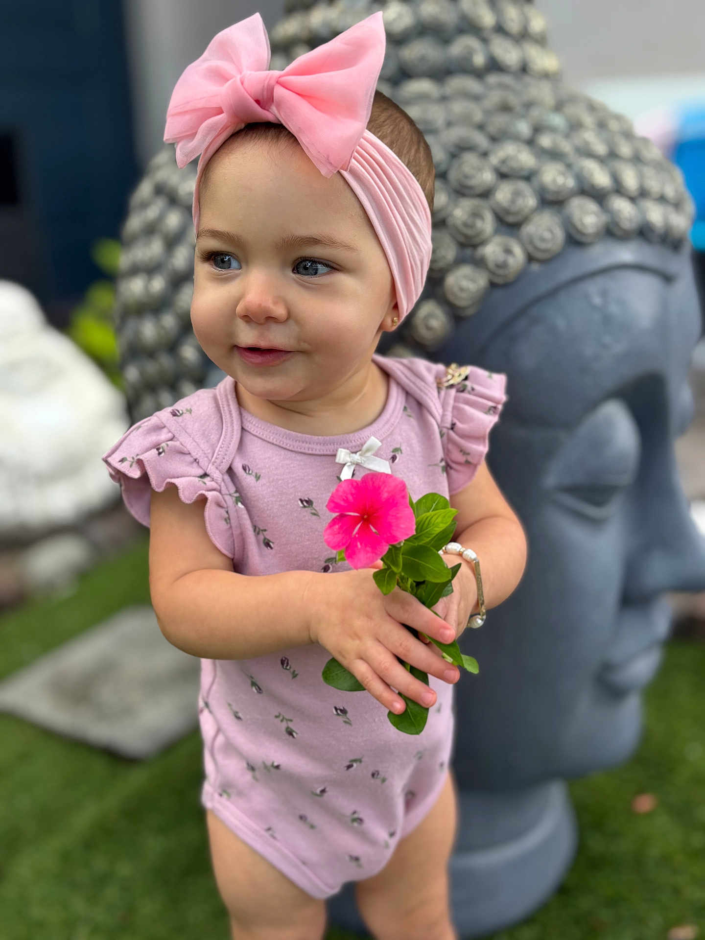Ainhoa joined the competition — help win amazing prizes! toddler, child, flower, pink_bow, pink_outfit, bracelet, buddha_statue, garden, greenery, grass, cute, portrait, smiling, headband, outdoor, person, nature, statue, happy, baby