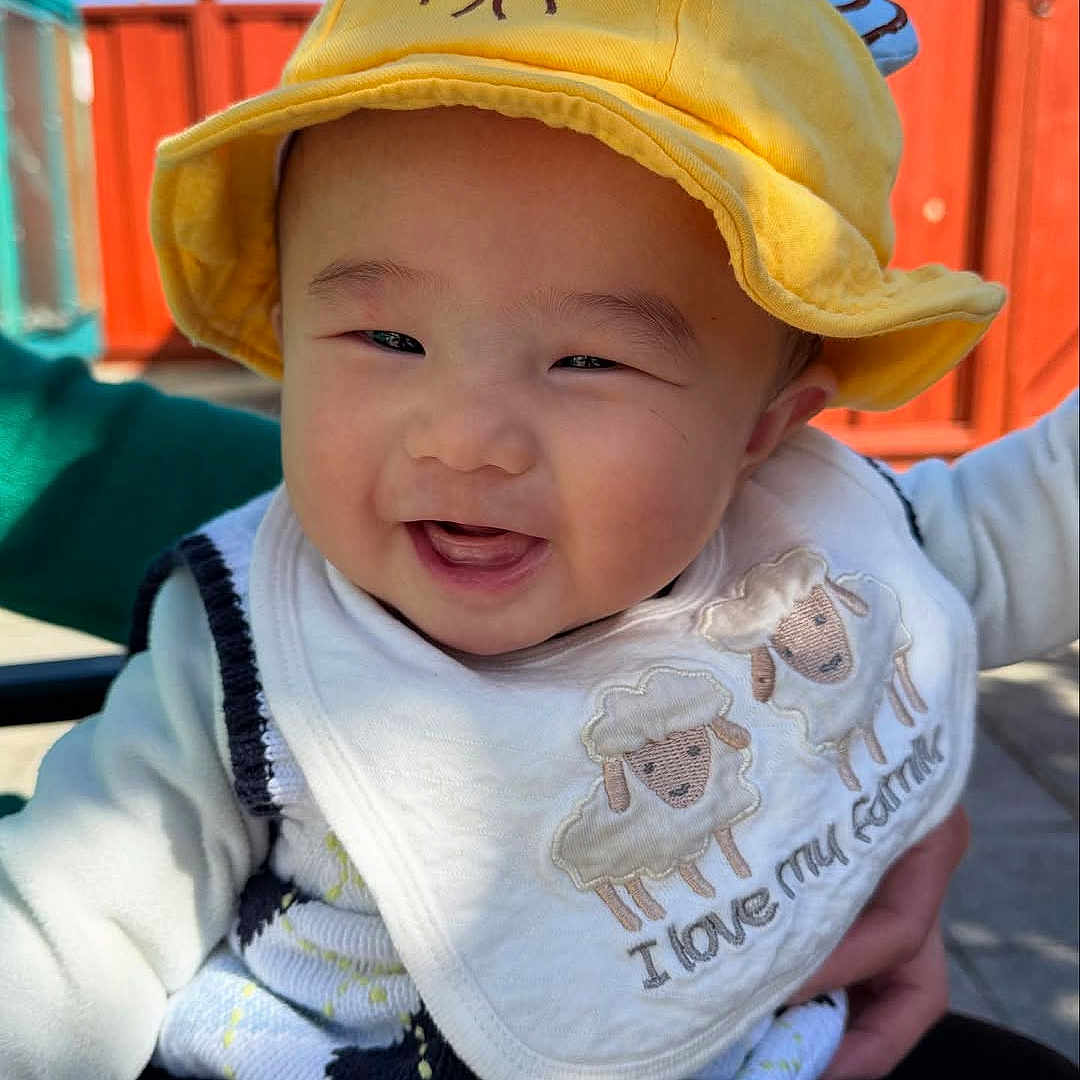Lucas is registered to the contest to win money with this photo: baby, background, bib, child, clothing, cute, daylight, embroidered_sheep, expression, face, happy, headwear, infant, outdoor, person, portrait, red_fence, smiling, sunny, yellow_hat