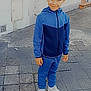 child, boy, smile, blue_tracksuit, white_sneakers, pavement, outdoor, sidewalk, urban, building, window, brick_wall, street, casual_clothing, standing, person, young, portrait, daylight, playful