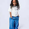 child, smiling, curly_hair, barefoot, blue_pants, white_top, standing, hands_in_pockets, happy, portrait, studio, fashion, casual, clothing, person, cute, young, full_length, isolated, background