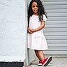 Kasey Gutierrez is registered to the contest to win money with this photo: child, girl, curly_hair, white_dress, red_polka_dots, red_sneakers, smiling, standing, leaning, wall, sidewalk, urban, outdoor, portrait, casual, happy, person, concrete, background, fashion