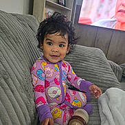 Elisa is registered to the contest to win money with this photo: toddler, child, baby, portrait, sitting, smiling, indoors, couch, blanket, pajamas, purple_clothing, donut_pattern, curly_hair, earrings, bottle, television, living_room, furniture, cozy, family_home