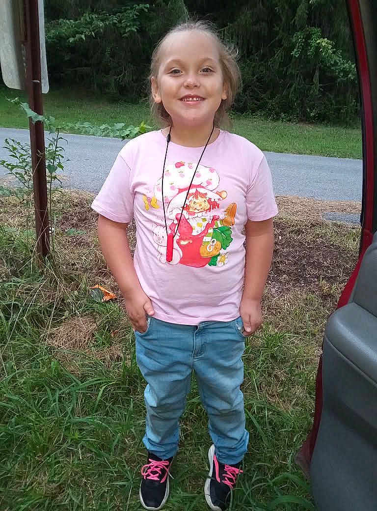 Rose is registered to the contest to win money with this photo: child, smiling, outdoor, grass, jeans, tshirt, sneakers, road, trees, car, person, nature, happy, casual_clothing, footwear, standing, greenery, portrait, daylight, accessory