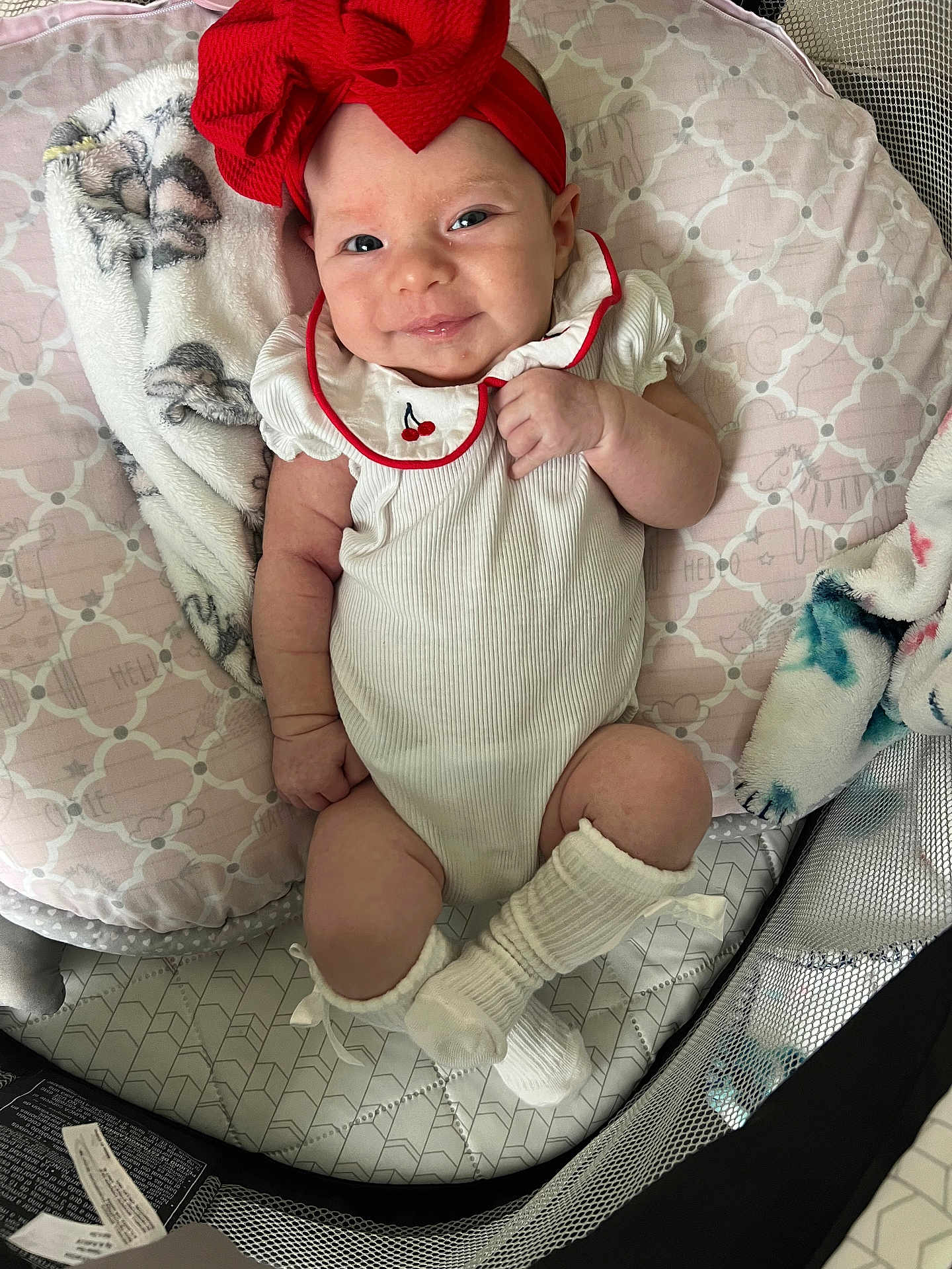 Charlee is registered to the contest to win money with this photo: baby, infant, smiling, red_bow, headband, white_outfit, blanket, cushion, soft_toys, lying_down, indoor, cute, child, happy, person, sock, playpen, patterned, cozy, portrait