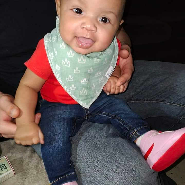 Nevaeh is registered to the contest to win money with this photo: baby, child, smiling, sitting, indoor, red_shirt, blue_jeans, pink_shoes, green_bib, person_holding, hands, happy, cute, infant, denim, footwear, casual_clothing, portrait, close_up, family
