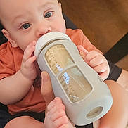 Leo is registered to the contest to win money with this photo: baby, bottle, feeding, infant, foot, hand, skin, person, indoor, closeup, cute, child, blue_eyes, drinking, holding, sitting, casual, warm_tone, adult_hand, floor