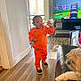toddler, child, baby_bottle, orange_clothing, sneakers, dog, couch, television, video_game, wooden_floor, indoor, person, pet, living_room, window, door, natural_light, flooring, furniture, casual