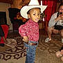 Chosen is registered to the contest to win money with this photo: child, cowboy_hat, red_shirt, jeans, belt, boots, living_room, carpet, curtains, wooden_walls, lamp, plant, woman, phone, sitting, photography, portrait, indoor, person, home
