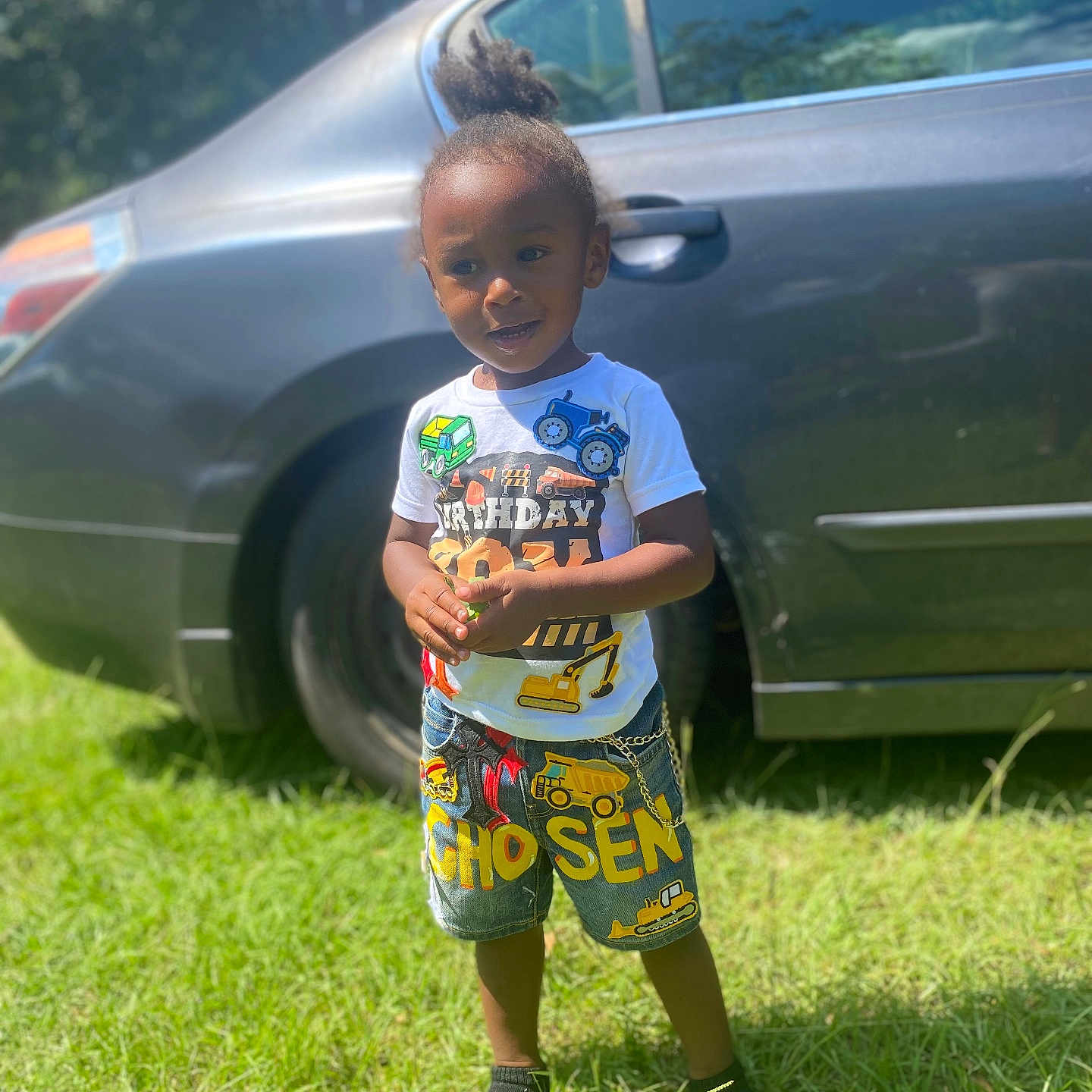 Chosen is registered to the contest to win money with this photo: car, casual_clothing, child, construction_vehicles, daylight, footwear, grass, greenery, hair_tied_up, happy, outdoor, parked_car, patches, person, shorts, smiling, standing, sunlight, toddler, tshirt