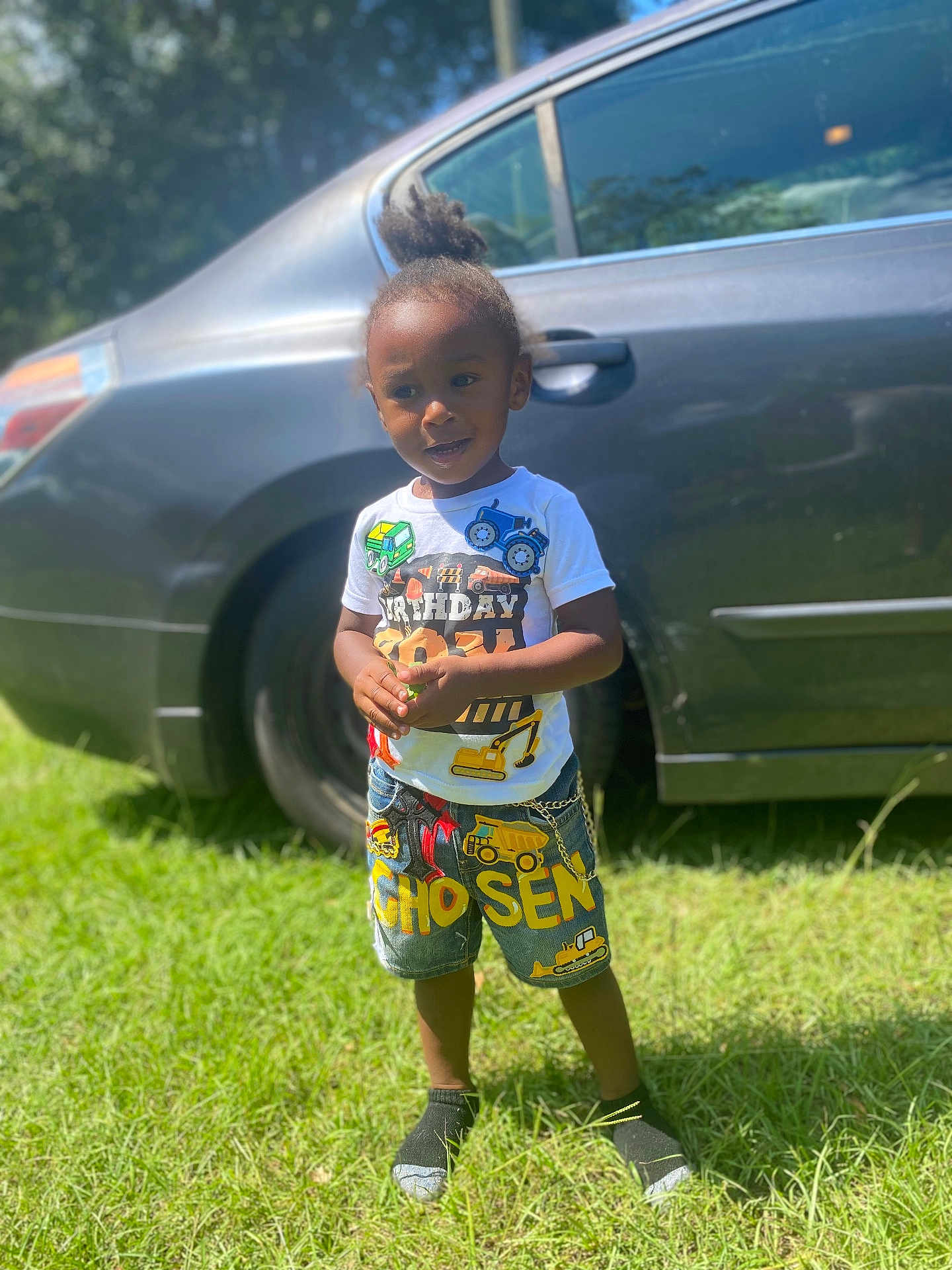Chosen is registered to the contest to win money with this photo: toddler, child, grass, car, outdoor, sunlight, shorts, tshirt, construction_vehicles, patches, smiling, standing, greenery, person, hair_tied_up, daylight, casual_clothing, footwear, happy, parked_car