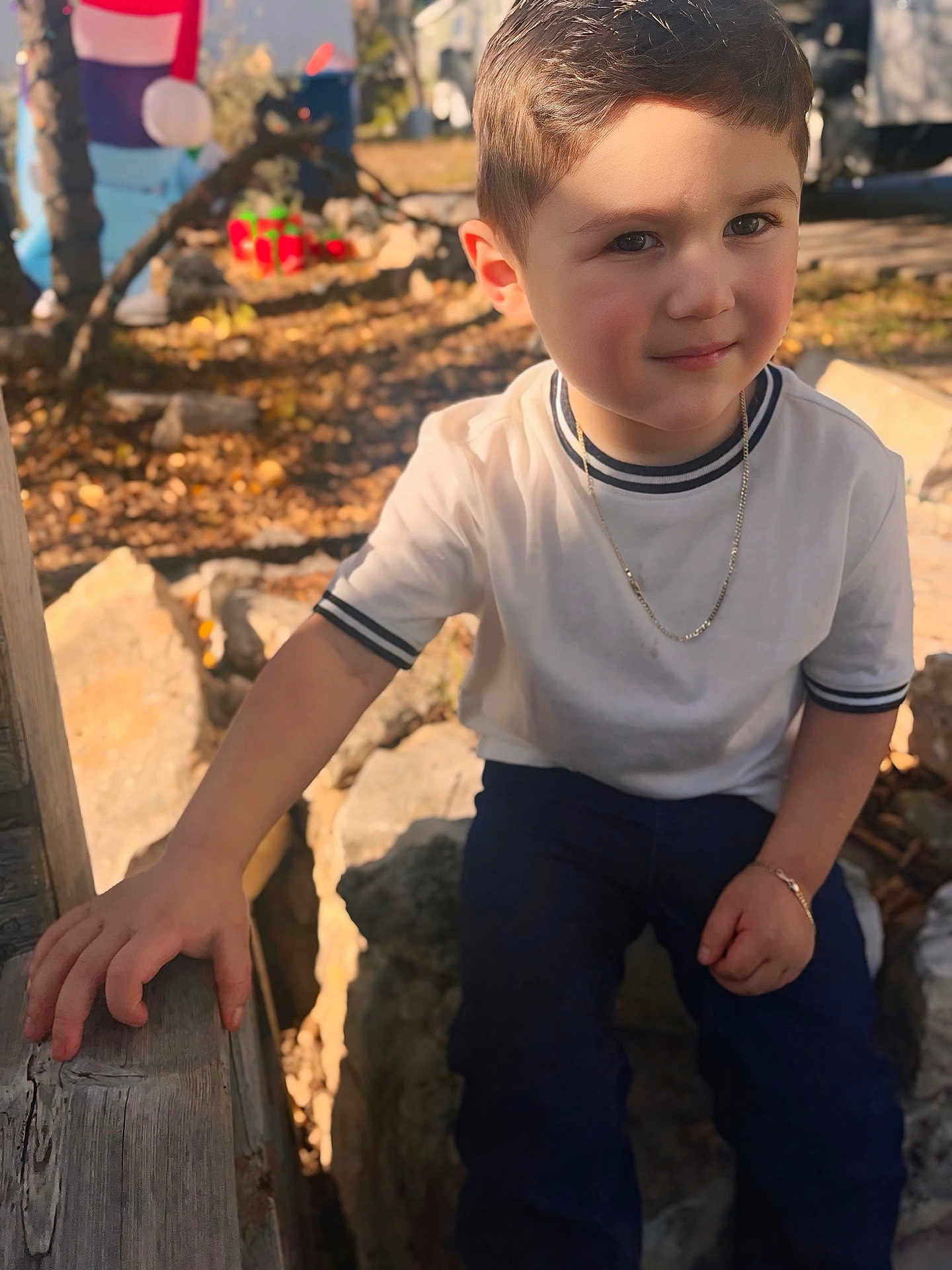 Domingo is registered to the contest to win money with this photo: child, boy, portrait, smile, face, chain_necklace, bracelet, white_shirt, jeans, hand, wooden_rail, rocks, leaves, autumn, sunlight, shadow, sitting, outdoors, close_up, short_hair