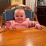 Ceaira is registered to the contest to win money with this photo: baby, child, high_chair, pink_sweatshirt, nike, wooden_chair, cushion, smiling, happy, indoor, kitchen, table, hand, face, person, photography, framed_photo, refrigerator, cabinet, light_switch