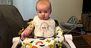 Kaisyn is registered to the contest to win money with this photo: baby, child, infant, bib, fisher_price, activity_seat, toy, colorful, indoor, living_room, couch, blanket, floor, wood_floor, television, cabinet, curious, seated, footwear, home