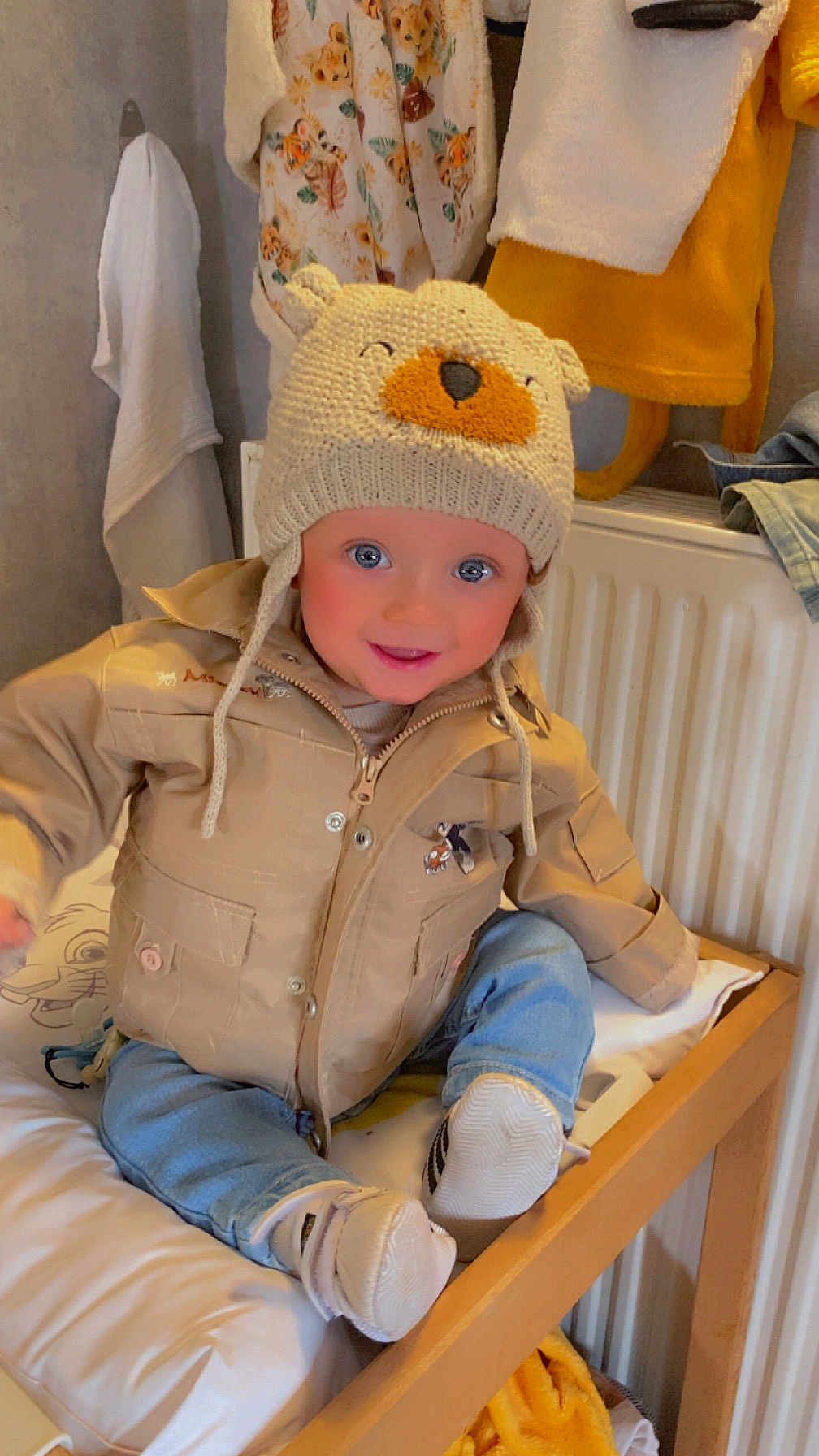 Eden participe au concours pour gagner de l'argent avec cette photo : toddler, child, hat, knitted_hat, jacket, beige_jacket, blue_jeans, shoes, smiling, indoor, changing_table, blanket, clothing, face, person, cute, warm_lighting, animal_print, home, baby