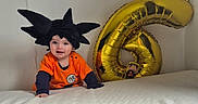 Elyas participe au concours pour gagner de l'argent avec cette photo : toddler, child, costume, wig, orange_clothing, balloon, gold_balloon, number_six, bed, white_bedspread, toy_figure, naruto_figure, indoor, celebration, happy, smiling, crawling, cute, birthday, party