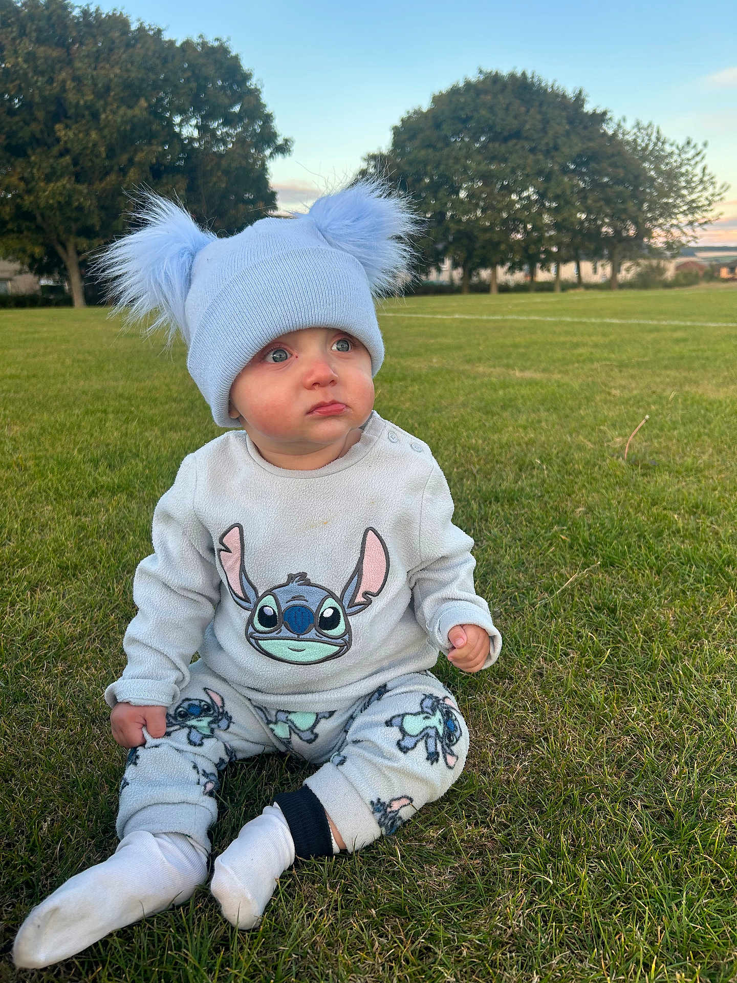 Arlo joined the competition — help win amazing prizes! baby, child, grass, outdoor, hat, blue_hat, pajamas, cartoon_character, sitting, cute, winter_clothing, pom_poms, green_grass, trees, nature, daylight, expression, curious, pout, portrait