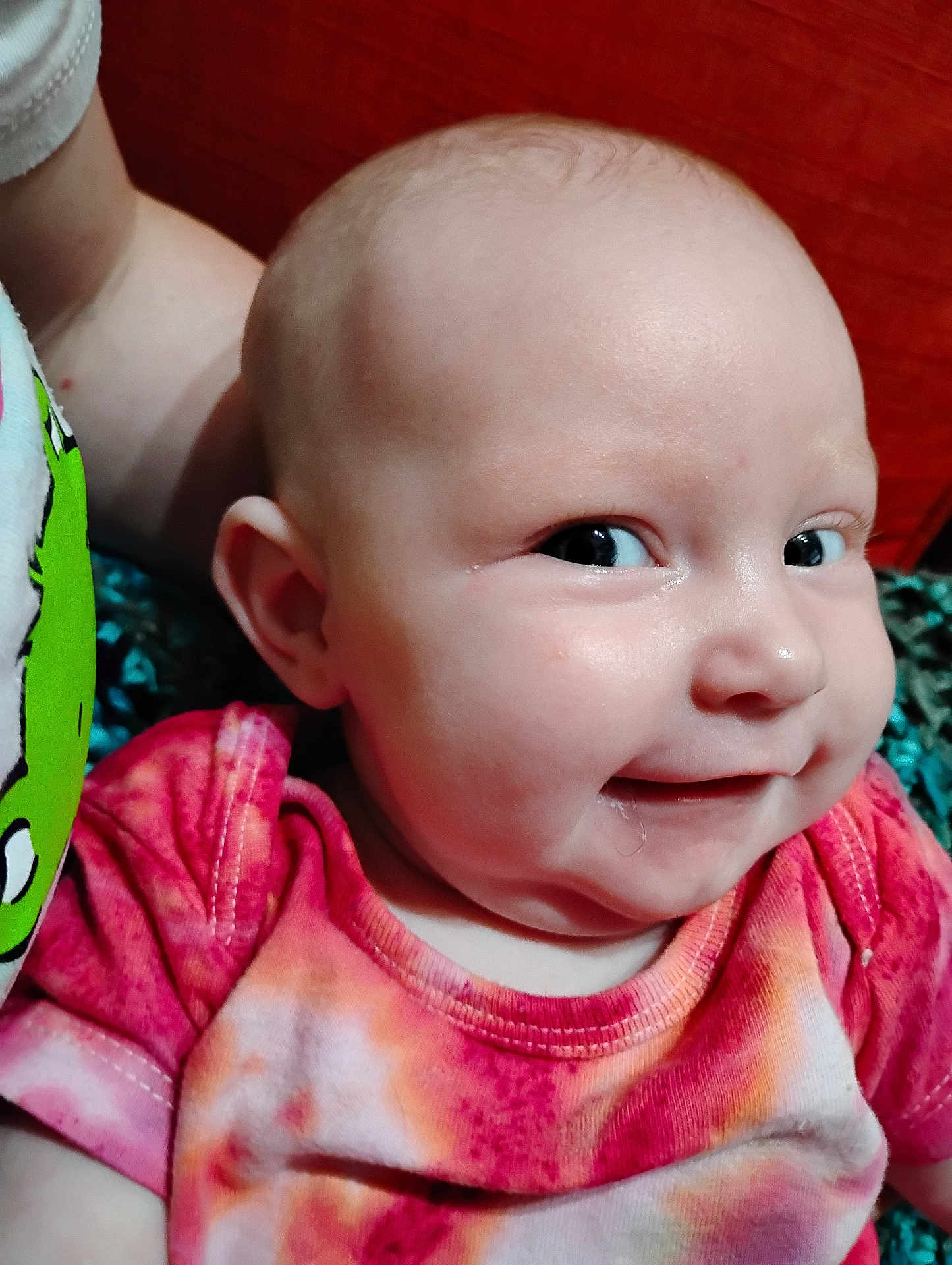 Macie is registered to the contest to win money with this photo: baby, infant, face, smile, cheeky, tie_dye, shirt, drool, close_up, person, child, skin, head, expression, cute, indoors, bright_eyes, cheek, colorful_clothing, playful