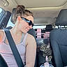 car_interior, casual, cat, center_console, cup_with_straw, curious, daylight, driver_seat, earrings, hair_bun, headrest, pink_objects, seatbelt, smile, sunglasses, tabby_cat, tank_top, tattoo, window, woman