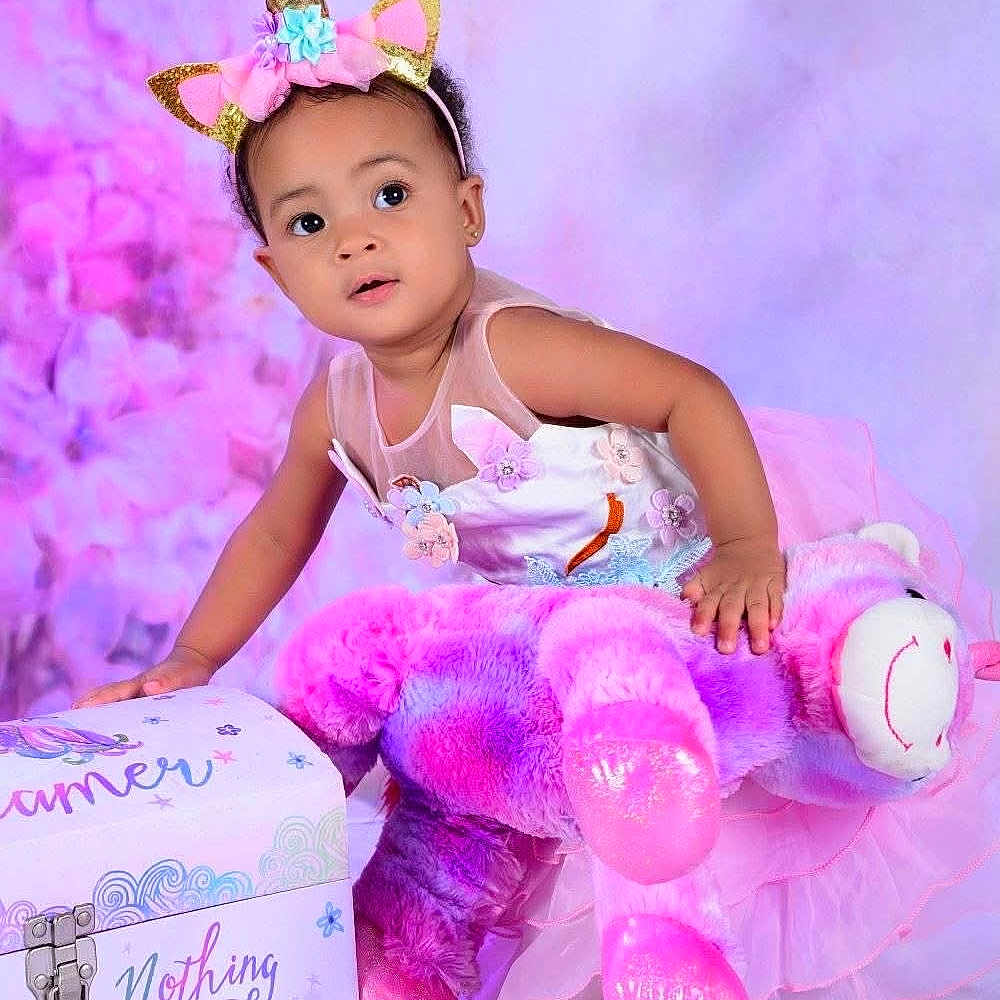Lovely is registered to the contest to win money with this photo: baby, child, curious_expression, cute, decorative_box, dress, flower_details, indoor, pastel_colors, pink, plush_toy, portrait, purple, smiling, soft_lighting, studio_background, teddy_bear, toddler, unicorn_headband, white_shoes