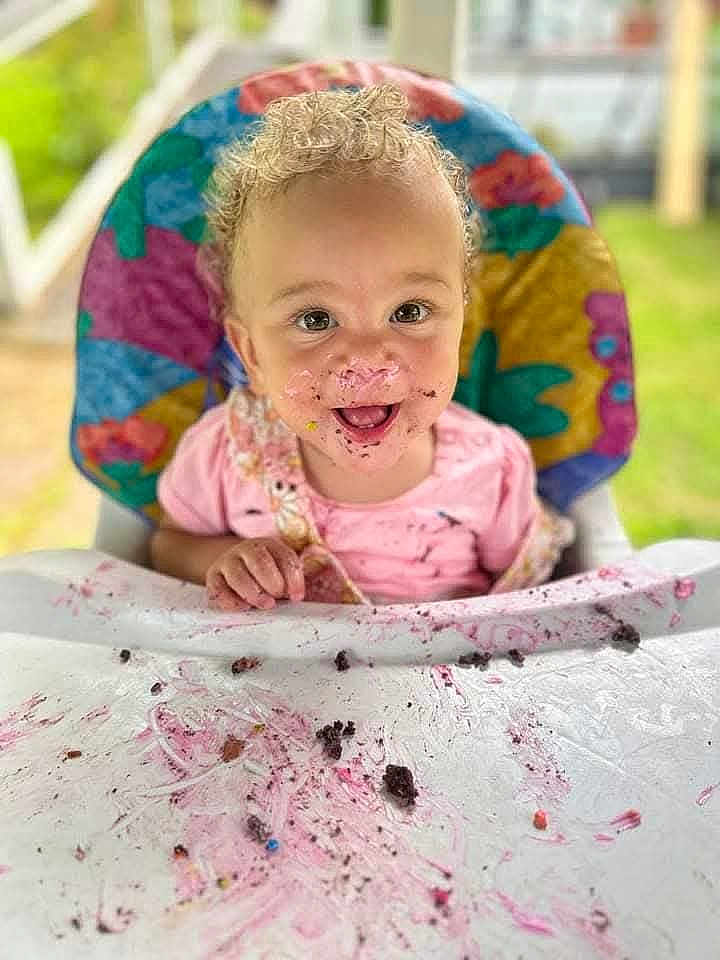 Isabella is registered to the contest to win money with this photo: toddler, child, high_chair, curly_hair, smiling, messy, cake, dessert, food, face_paint, happy, indoors, colorful, table, seat, playful, young_child, portrait, celebration, fun