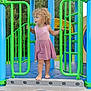 child, toddler, playground, barefoot, curly_hair, pink_dress, green_bars, blue_platform, outdoor, trees, summer, play_equipment, adventure, young_child, grass, daytime, nature, fun, exploration, park