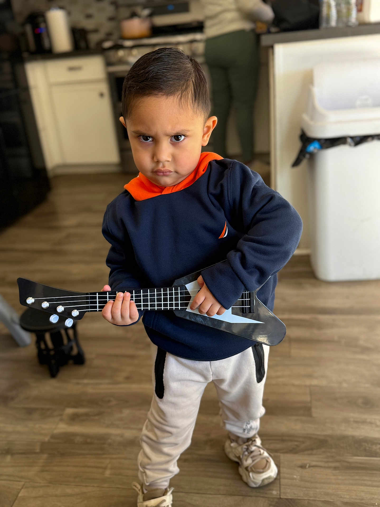 Henry is registered to the contest to win money with this photo: child, toddler, toy_guitar, serious_expression, indoor, wooden_floor, sneakers, casual_clothing, person, standing, kitchen, pants, sweatshirt, short_hair, playing, musical_instrument, young_child, focused, home, cute