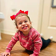 Emberlee is registered to the contest to win money with this photo: toddler, child, smiling, crawling, red_bow, pink_sweater, wooden_floor, indoors, happy, cute, person, face, hands, young_child, casual_clothing, playful, bright, home, cheerful, portrait
