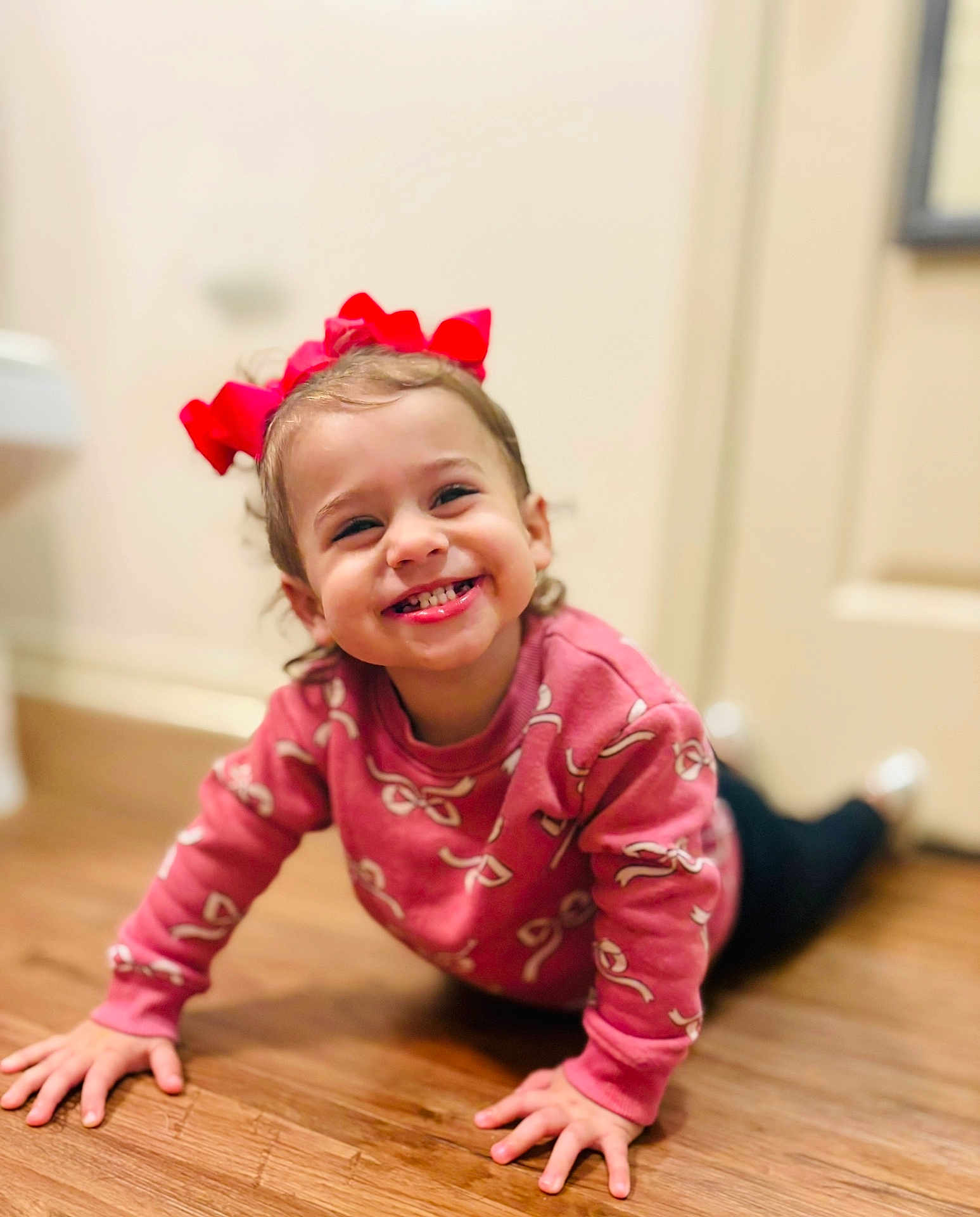 Emberlee is registered to the contest to win money with this photo: toddler, child, smiling, crawling, red_bow, pink_sweater, wooden_floor, indoors, happy, cute, person, face, hands, young_child, casual_clothing, playful, bright, home, cheerful, portrait