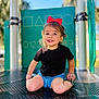 toddler, child, red_bow, black_shirt, blue_shorts, playground, outdoor, smiling, happy, sitting, metal_surface, daylight, nature, trees, fun, cute, young_child, person, play, casual_clothing