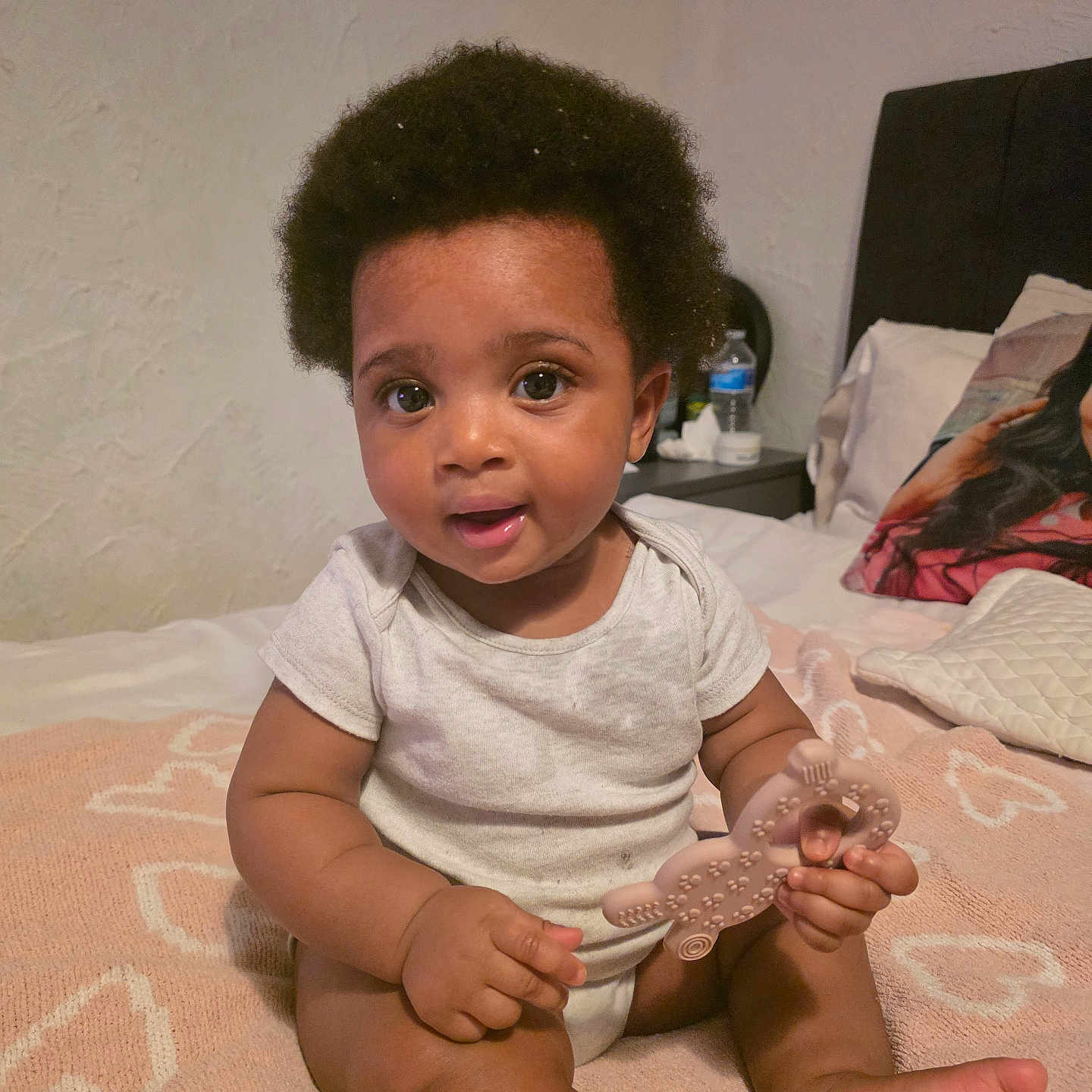 Kyla joined the competition — help win amazing prizes! afro, baby, bed, blanket, child, cute, foot, hand, heart_pattern, indoors, infant, nightstand, pillow, portrait, sitting, skin, smiling, teether, water_bottle, white_onesie