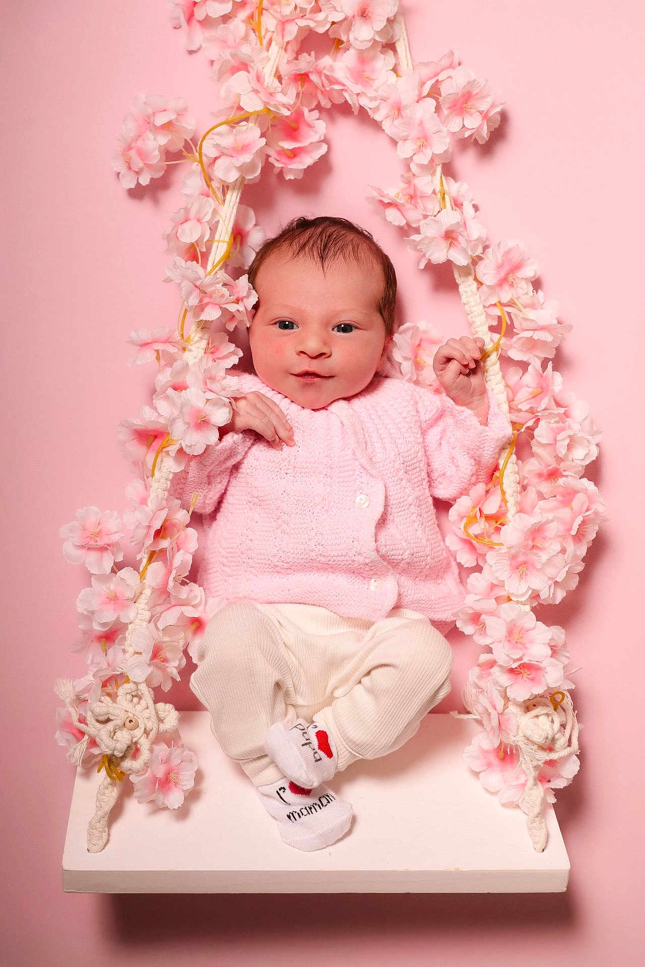 Ava participe au concours pour gagner de l'argent avec cette photo : baby, infant, pink, swing, flowers, cute, child, portrait, knitted_sweater, white_pants, socks, studio, background, flower_garland, smiling, seated, young_child, soft_lighting, indoors, decorative