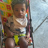 Kamryn is registered to the contest to win money with this photo: baby, bib, child, clothing, colorful, curly_hair, daylight, eyes, face, harness, outdoor, pavement, photograph, portrait, seated, serious, sidewalk, stroller, sunglasses, toddler
