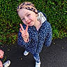 child, girl, smiling, peace_sign, headband, blue_jacket, floral_pattern, leggings, sneakers, outdoor, greenery, hedge, pavement, playful, portrait, casual_clothing, happy, fashion, nature, daylight