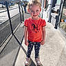 child, smiling, outdoor, sidewalk, sunlight, shadow, fence, street, building, window, door, orange_shirt, leggings, sneakers, person, daylight, happy, young, casual_clothing, urban