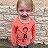 child, girl, smile, blonde_hair, orange_shirt, cartoon_print, leggings, sneakers, outdoor, stone_wall, standing, happy, casual_clothing, blue_eyes, portrait, daylight, concrete_ground, cute, young, person