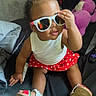toddler, child, sunglasses, red_skirt, polka_dot, white_top, sandals, indoor, plush_toy, purple_toy, cute, smiling, sitting, hand_on_face, hair_bun, playful, black_surface, casual, home, portrait