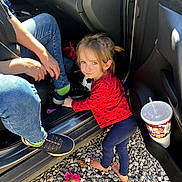 Cynthia is registered to the contest to win money with this photo: adult, barefoot, blue_pants, car, casual, child, cup, door, drink, flower, gravel, jeans, outdoor, person, red_shirt, smile, sneakers, sunlight, toddler, toy