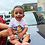 baby, building, car, child, clothing, face, foot, graffiti, hand, happy, outdoor, person, shorts, sitting, sky, smiling, support, tshirt, urban, vehicle